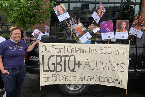 Monica Meyer, wearing one of Outfront Minnesota's "We The People means everyone" t-shirts, stands beside a banner which reads "OutFront Celebrates 50 Iconic LGBTQ+ Activists For 50 Years Since Stonewall," behind which photos of several of those icons are displayed on picket signs.