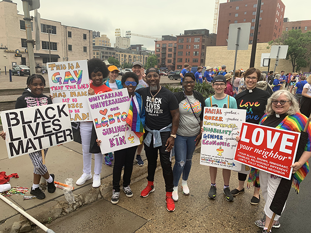 A small crowd stands with colorful shirts and signs reading slogans such as "Black Lives Matter," "Love they neighbor*," "Unitarian Universalists love our LGBTQ siblings and kin," and "Love is Love."