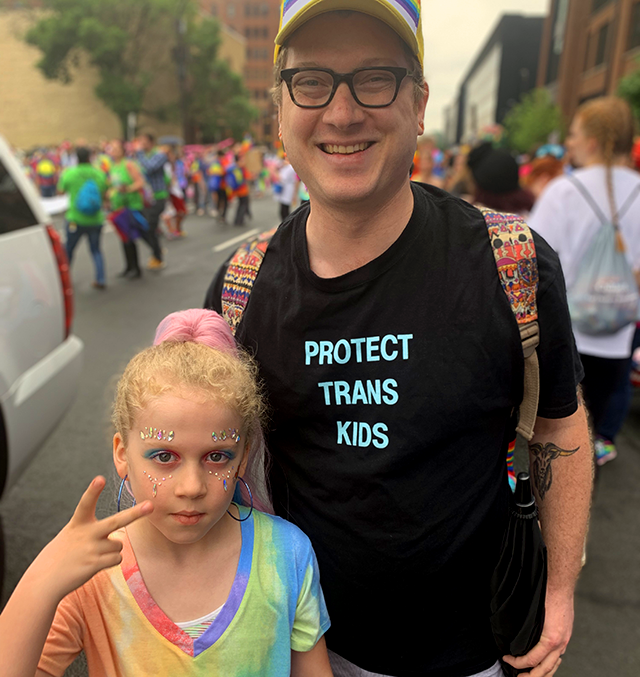 Dave and Hildie Edwards face the camera. Dave, wearing a shirt that reads "protect trans kids," smiles. Hildie, dressed in rainbow fashion, displays a peace sign to the camera.