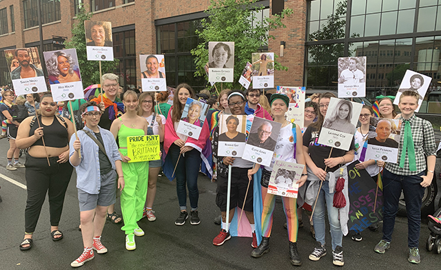 A small crowd stands together, each holding picket signs featuring influential figures in the LGBTQ+ community.