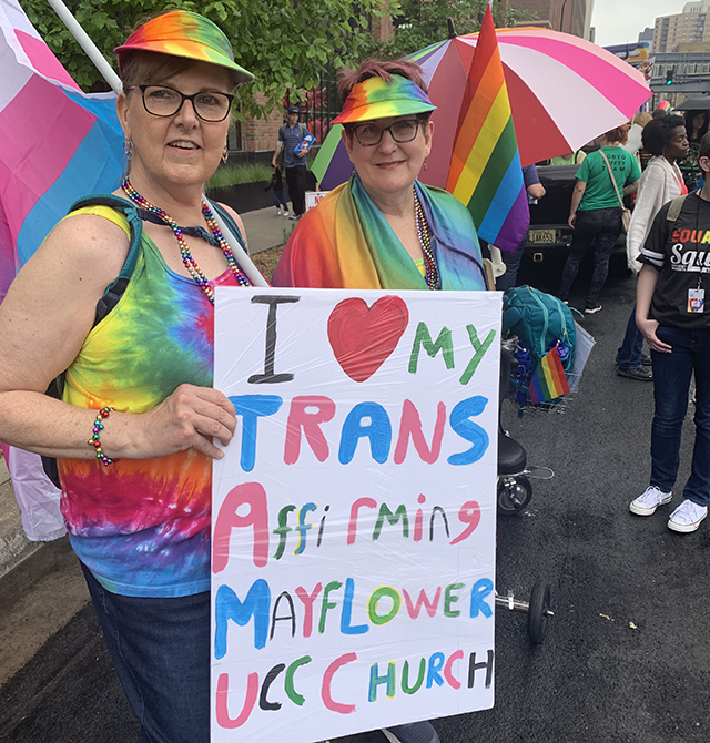 Stephanie and Carla Peck smile towards the camera. Dressed in rainbows and holding a transgender pride flag, they display a sign that reads "I love my trans affirming Mayflower UCC Church."
