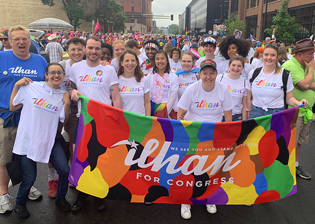 Supporters of Ilhan Omar, each wearing a shirt featuring her name written in a scrawling rainbow font, stand together holding a banner covered in colorful patterns and reading "We see you and stand with you" and "Ilhan for congress."