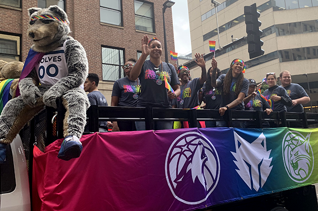 A float with a rainbow banner along its side featuring the logos of sports teams such as the Minnesota Timberwolves and Minnesota Lynx carries several waving individuals. The Timberwolves mascot, bearing a rainbow flag and headband, sits at the edge of the vehicle.