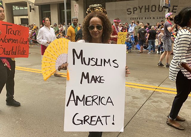 Leila Ali displays her "Muslims make America great!" sign to the camera while holding a yellow folding fan. Another poster reading "Trans women of color started pride" is visible in the background among the marchers as well.